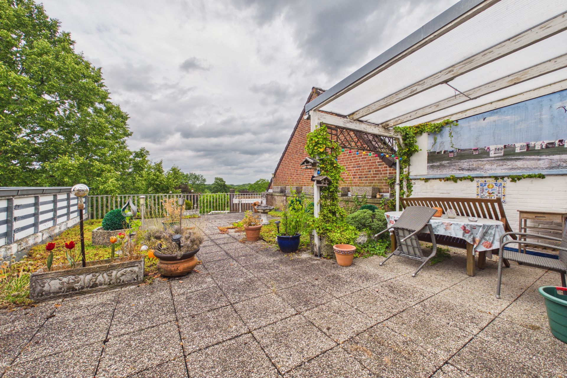 Große Terrasse mit Pflanzen, Sitzbank, Tisch und Überdachung, Blick ins Grüne.