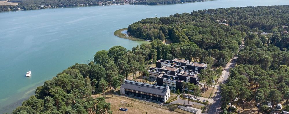 Luftaufnahme eines Gebäudekomplexes am Waldrand direkt an einem großen See.