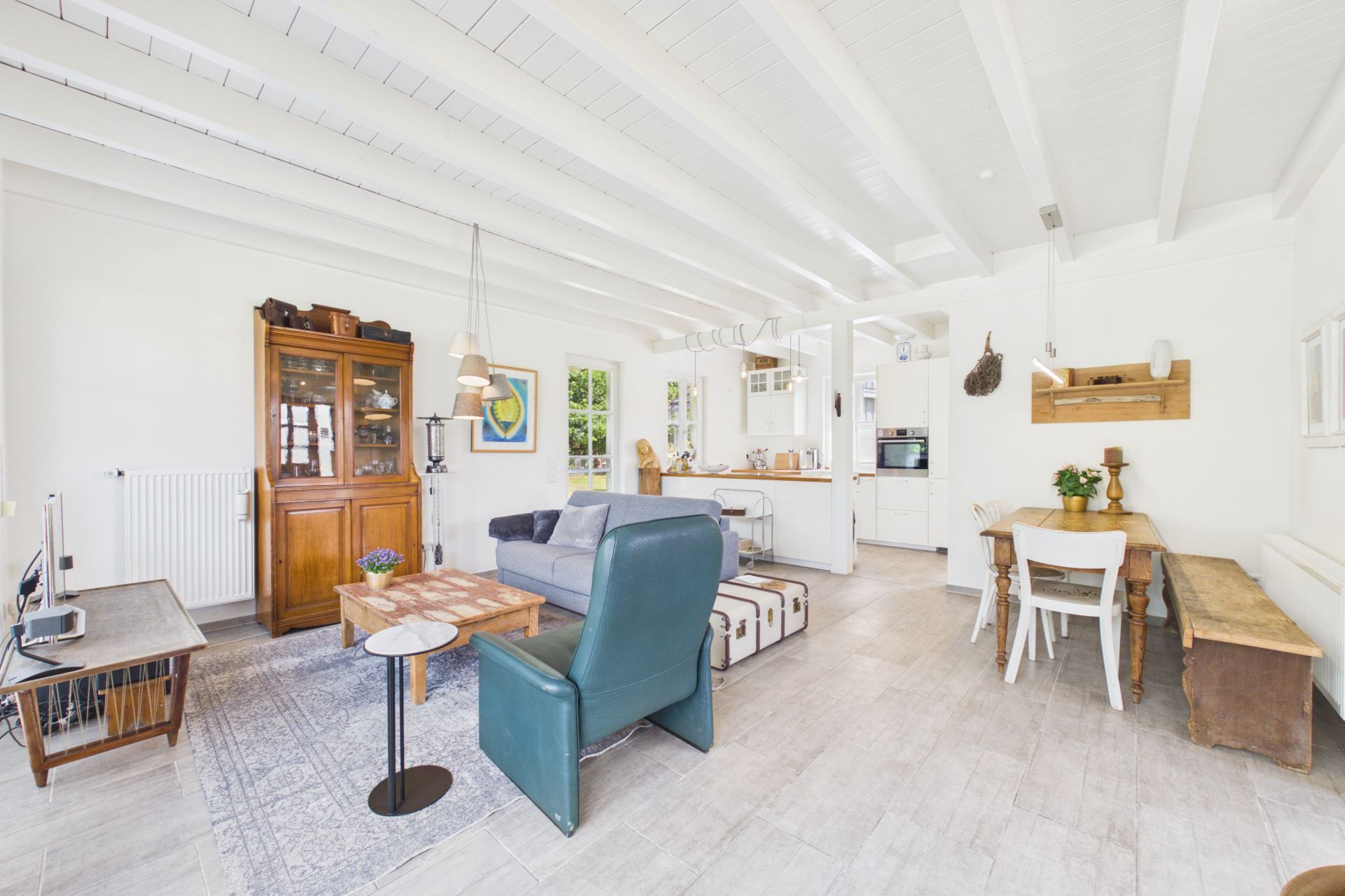 Helles Wohnzimmer mit Sofa, Sessel, Esstisch und offener Küche, Holzmöbeln und sichtbaren Deckenbalken.