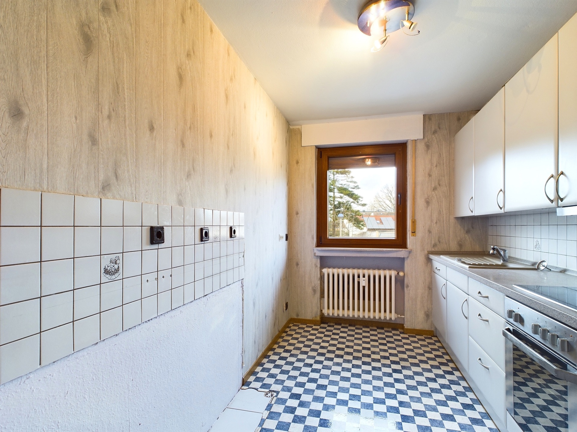 Schmale Küche mit weißen Schränken, kariertem Boden und Fenster am Ende des Raumes.