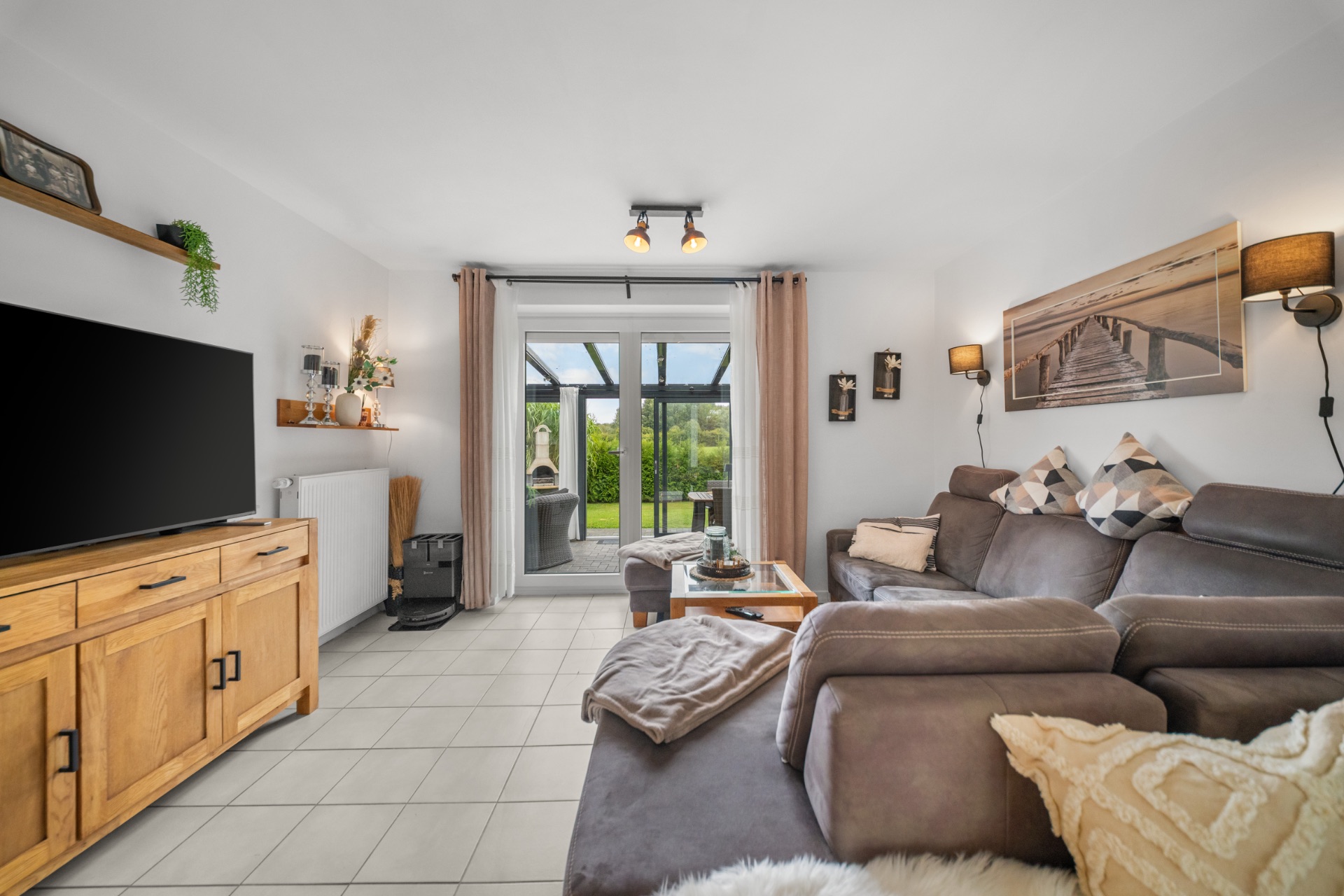 Helles Wohnzimmer mit grauem Sofa, Fernseher, Holzmöbeln und Zugang zur Terrasse.