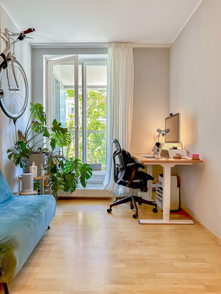 Helles Zimmer mit Schreibtisch, Stuhl, Sofa, Pflanze, Wandfahrrad und großem Fenster.