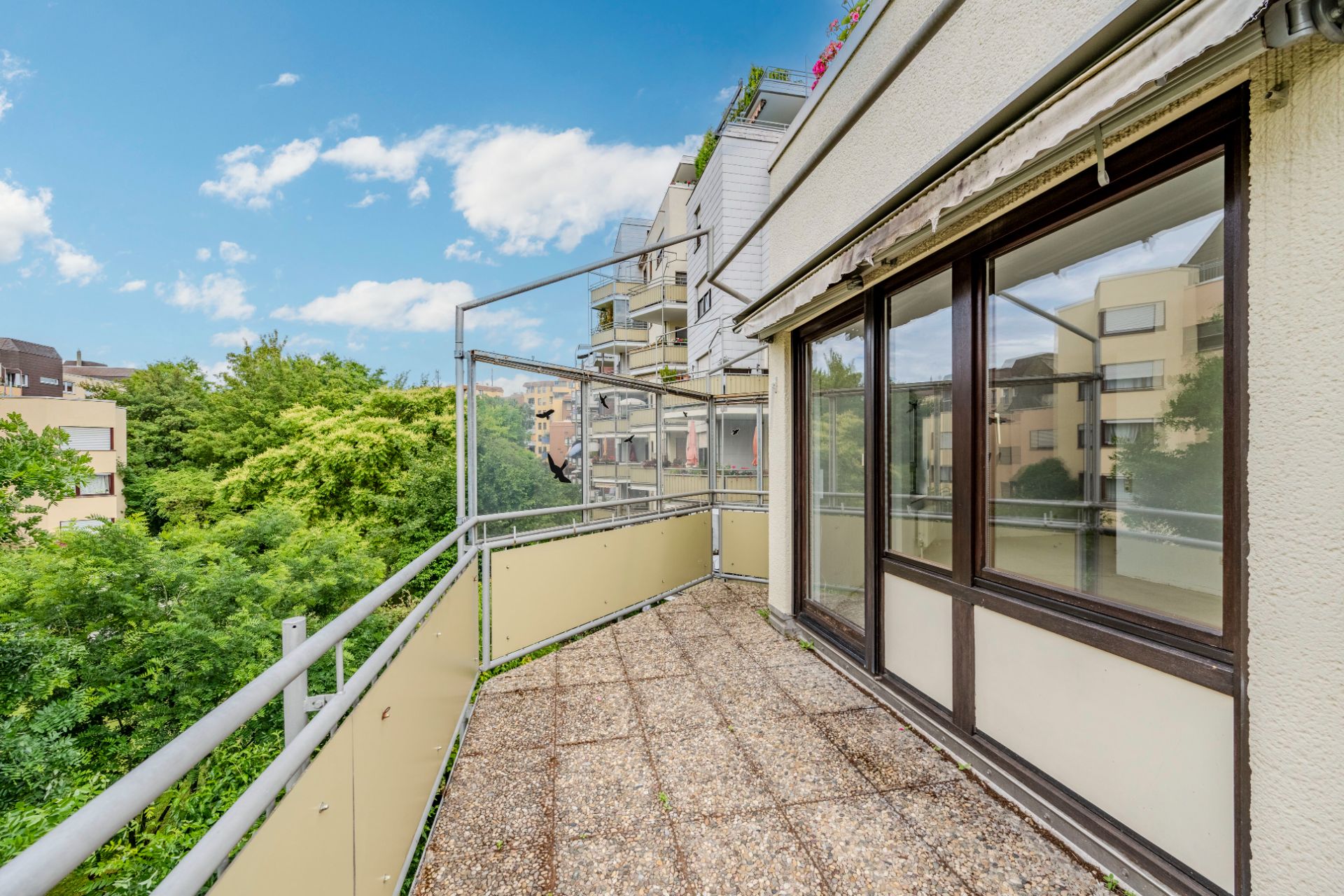 Balkon mit Geländer, Blick auf Bäume und angrenzende Wohngebäude bei blauem Himmel.