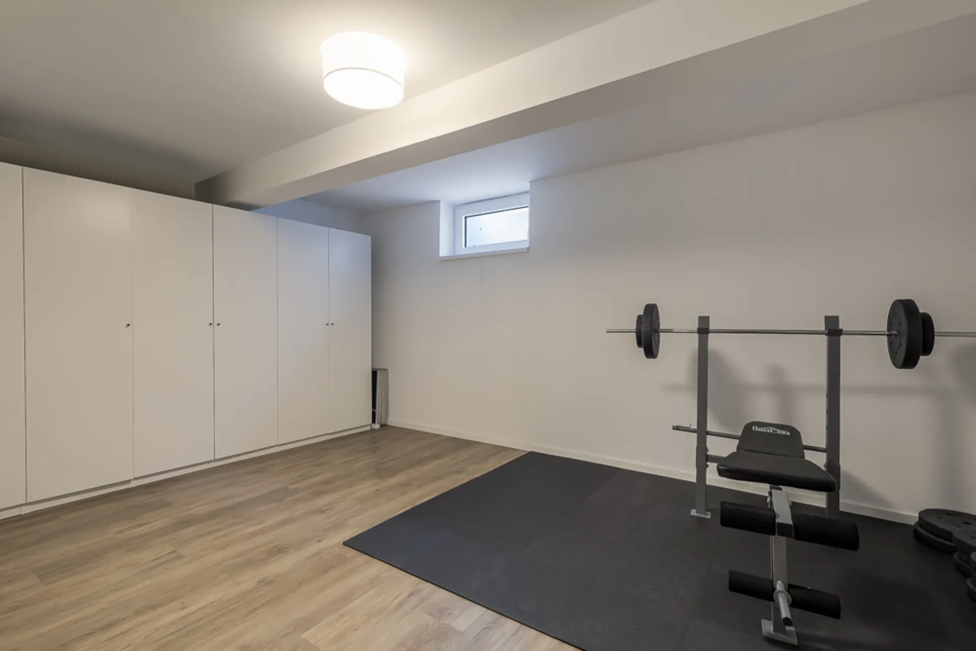 Raum mit weißen Schränken, einer Hantelbank und kleinem Fenster, Holzboden und Sportmatte.