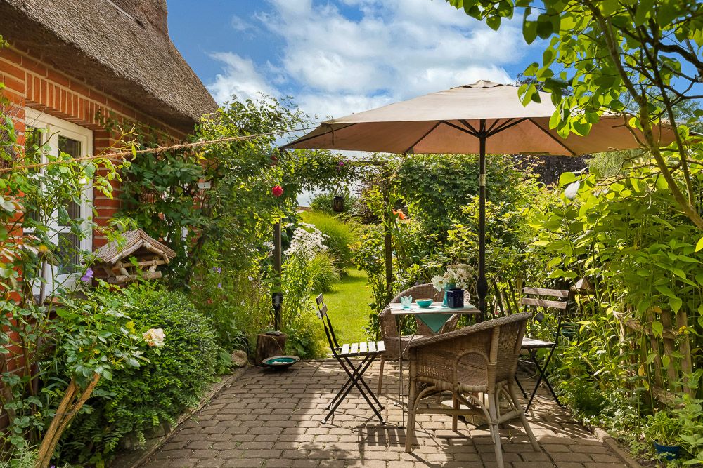 Gepflasterte Terrasse mit Tisch, Stühlen und Sonnenschirm in grünem Garten neben einem Ziegelhaus.
