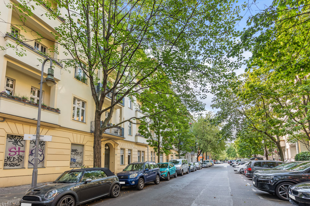 Baumgesäumte Wohnstraße mit parkenden Autos und mehrstöckigen Wohnhäusern.