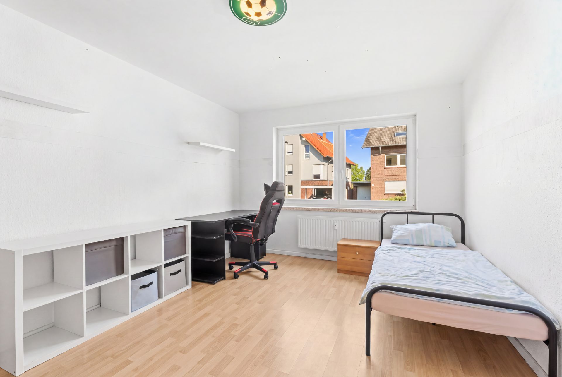 Helles Zimmer mit Einzelbett, Schreibtisch, Regal und Stuhl, Fenster mit Blick nach draußen.