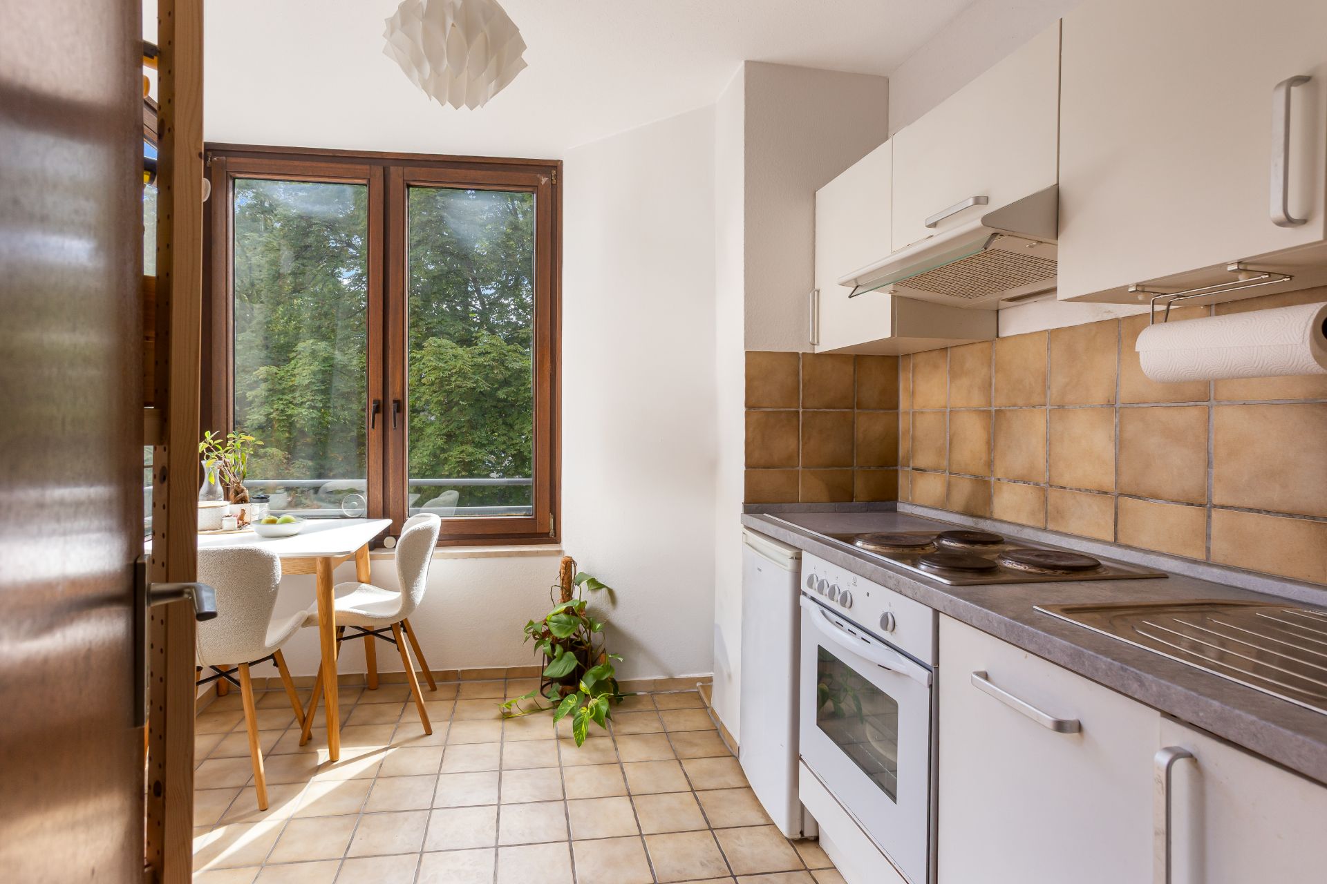 Helle Küche mit Esstisch, Herd, Spüle und großen Fenstern, eine Pflanze steht in der Ecke.