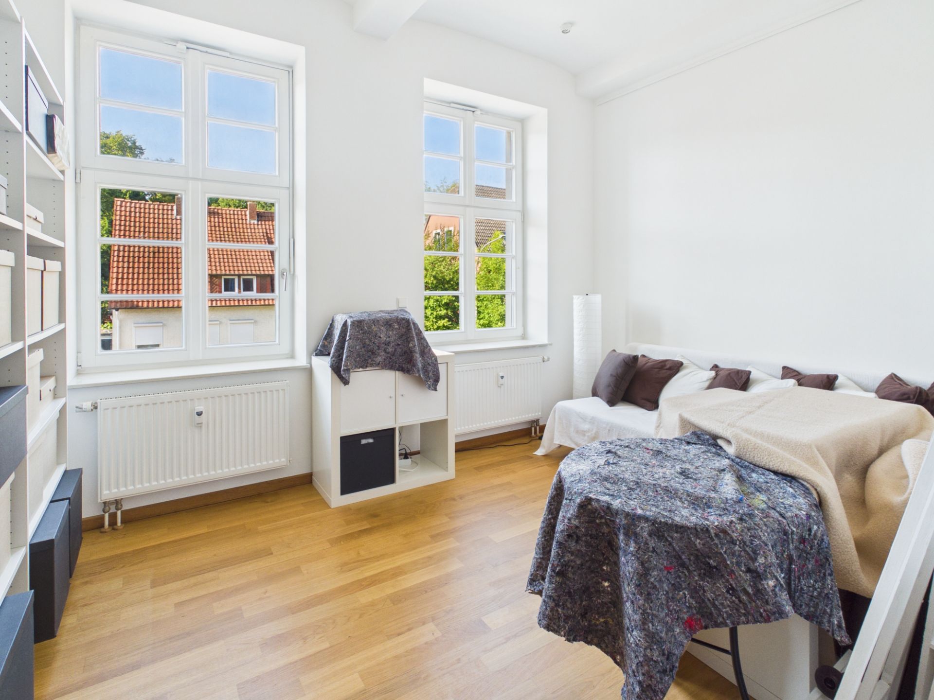 Helles Zimmer mit Holzboden, zwei Fenstern, Regal, Sofa und Möbeln, die mit Decken abgedeckt sind.