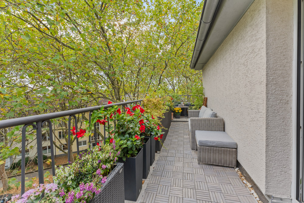 Langer Balkon mit Sitzmöbeln und vielen Blumenpflanzen, Blick auf Bäume und Nachbargebäude.