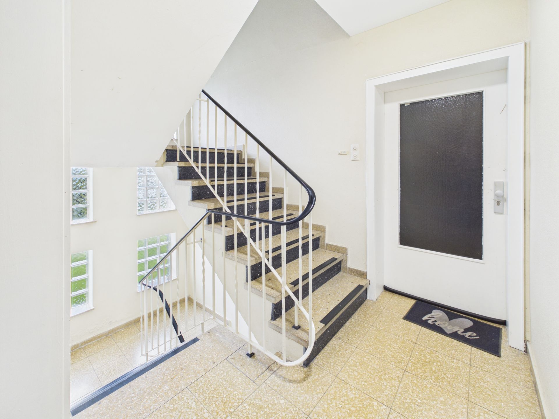 Helles Treppenhaus mit Geländer, Fenstern und weißer Wohnungstür mit Fußmatte.