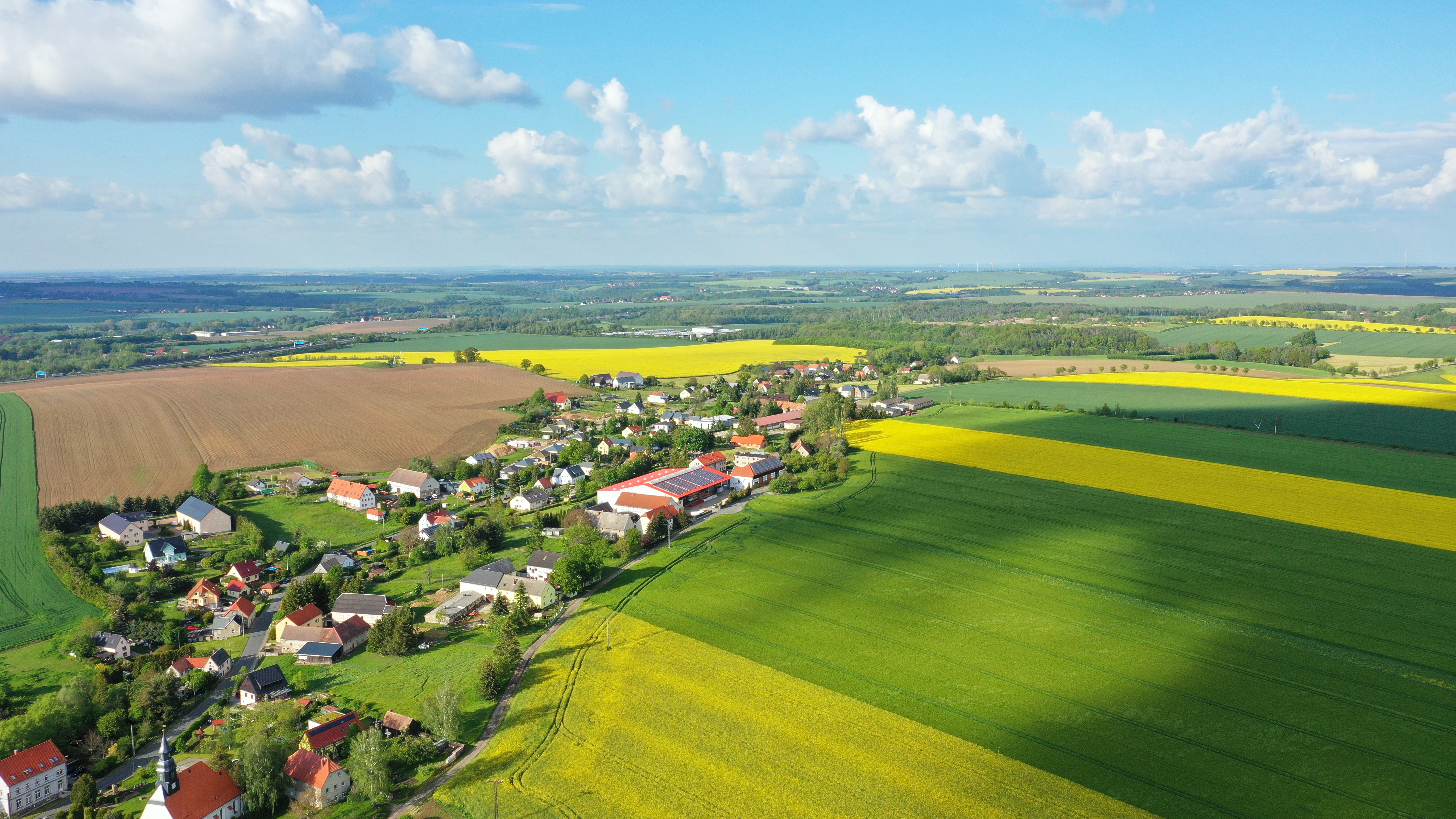 Luftaufnahme eines kleinen Dorfes mit umliegenden Feldern und Häusern bei Tageslicht.