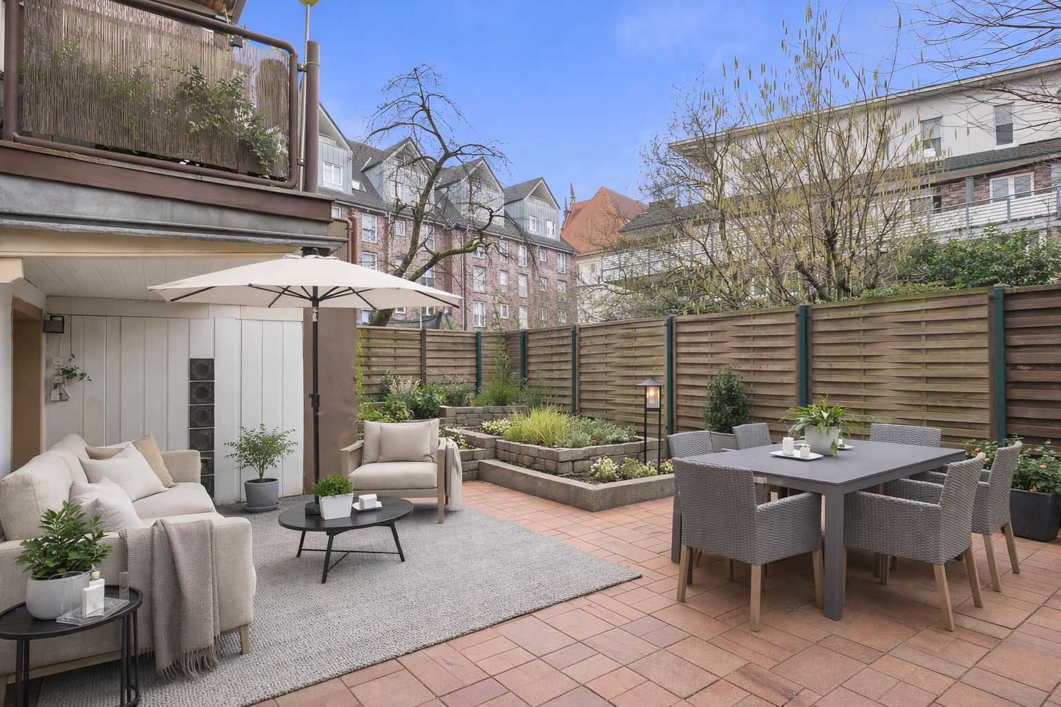 Moderne Terrasse mit Sofa, Gartentisch, Stühlen, Sonnenschirm und Pflanzen, umgeben von Holzzaun.