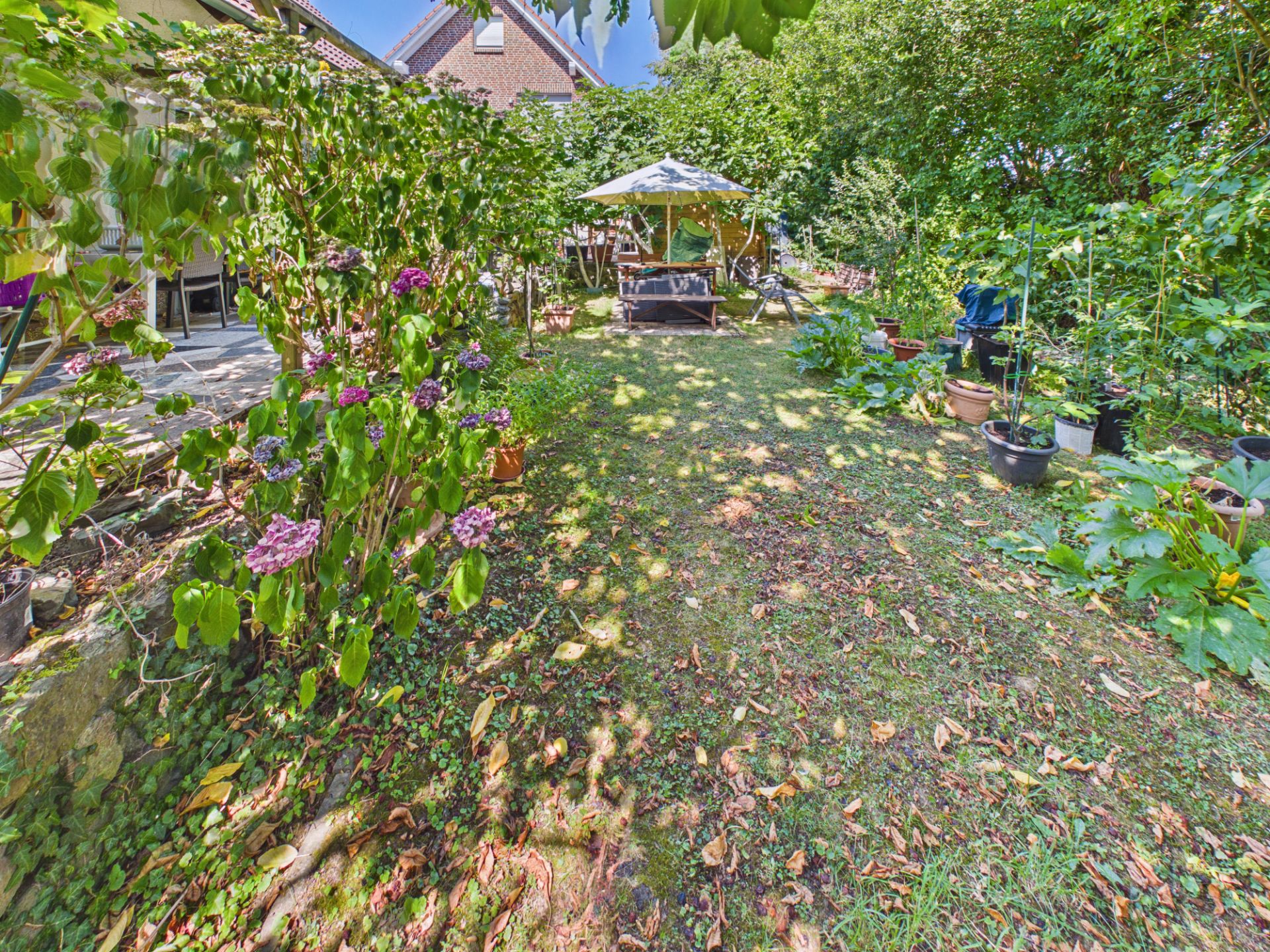 Grüner Garten mit Blumen, Bäumen und Sitzbereich unter einem Sonnenschirm.