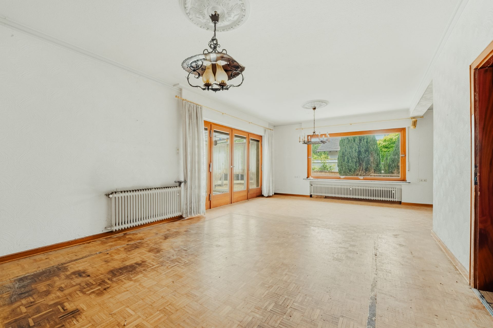 Geräumiges, leeres Wohnzimmer mit Parkettboden, großen Fenstern, Heizkörpern und zwei Deckenlampen.