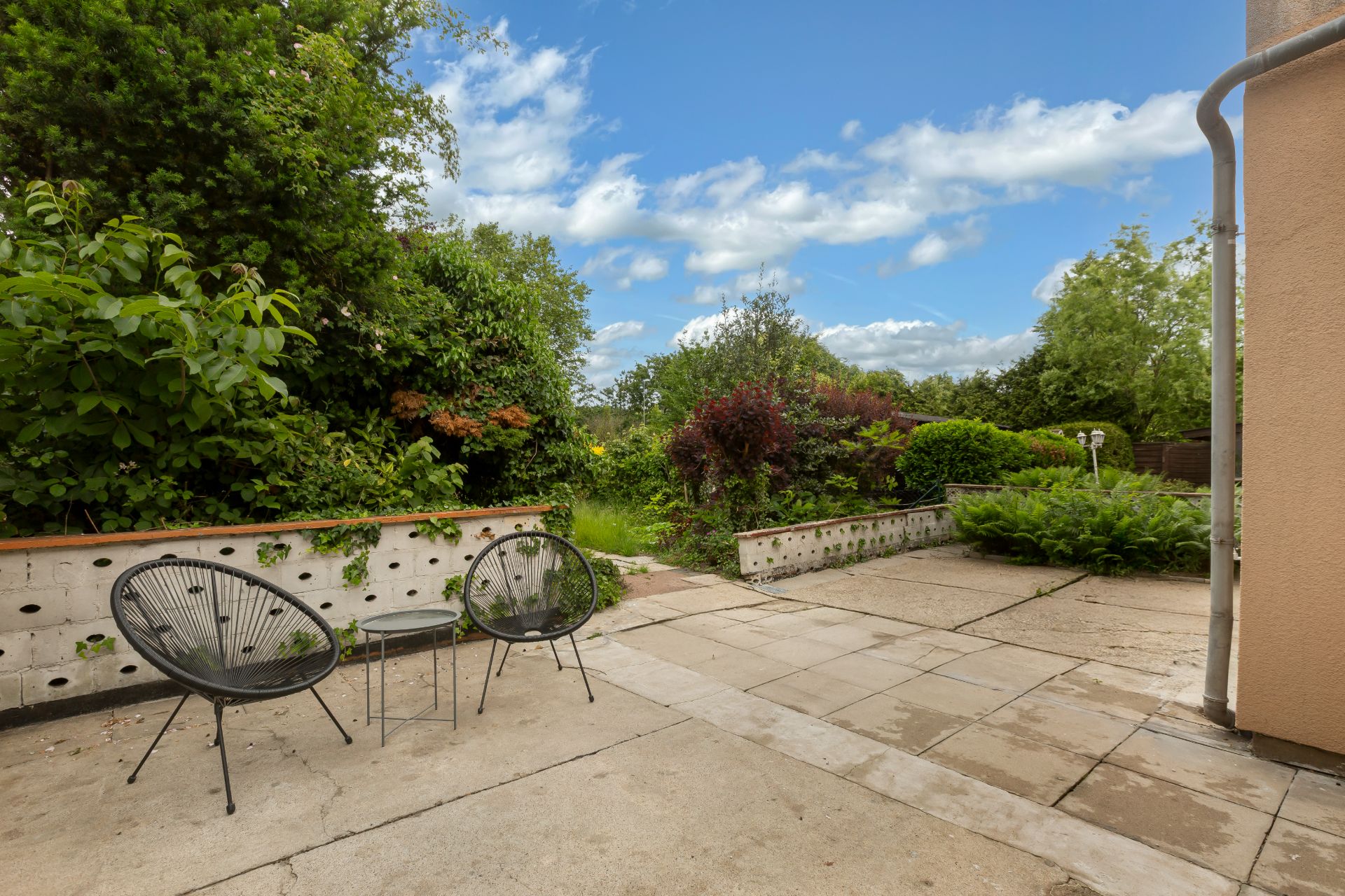 Gepflasterte Terrasse mit zwei Stühlen, kleinem Tisch und viel Grün im Garten.