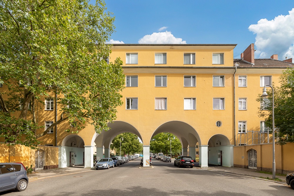 Gelbes Wohngebäude mit drei großen Rundbögen über einer Straße, geparkte Autos und Bäume daneben.