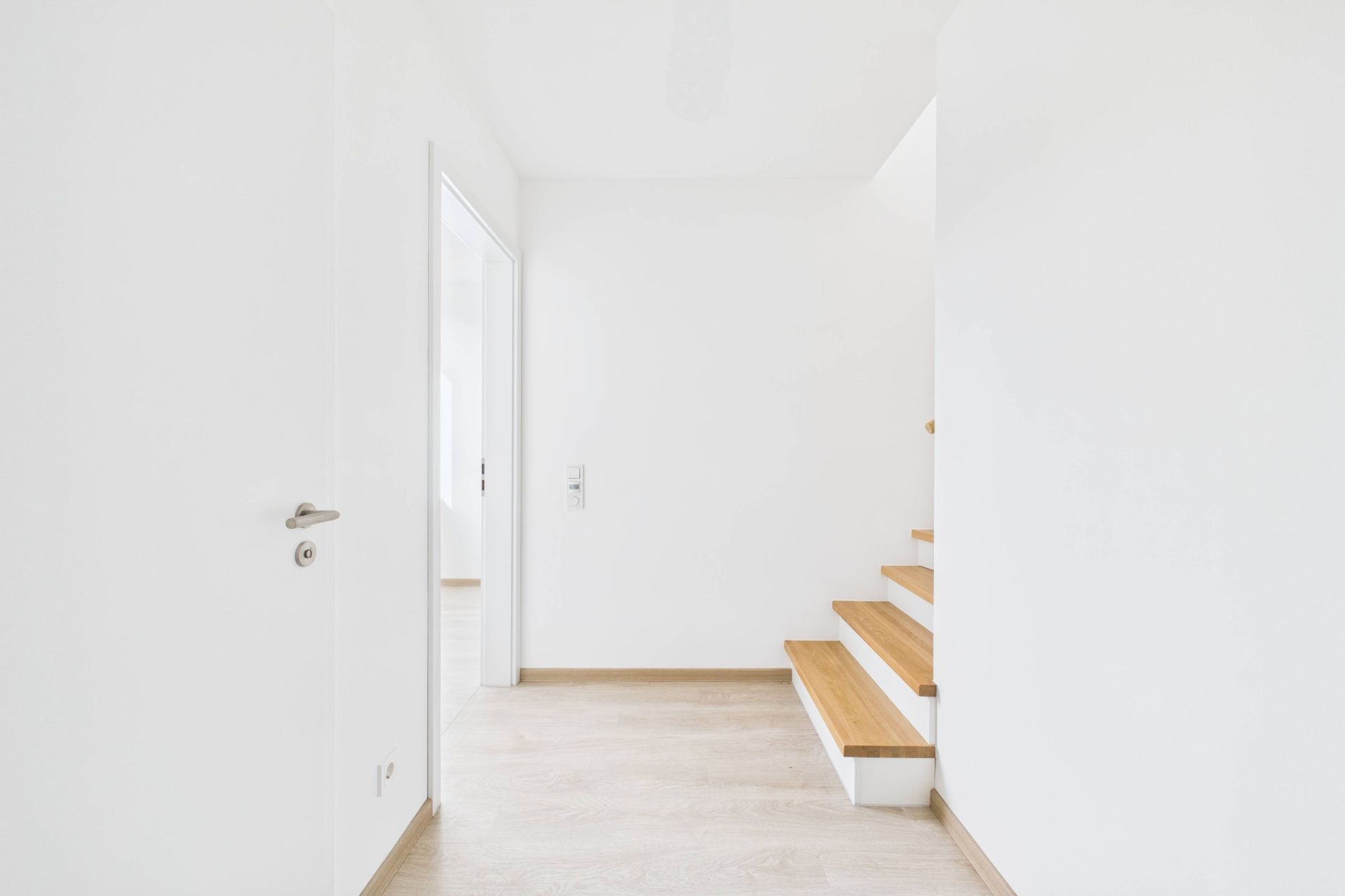 Heller Flur mit Holzboden, weißer Wand, Tür links und Treppe nach oben rechts.