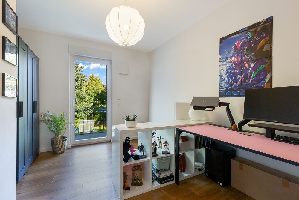 Helles Arbeitszimmer mit Schreibtisch, Regal, Computer, Pflanze und großen Fenstern mit Blick ins Grüne.