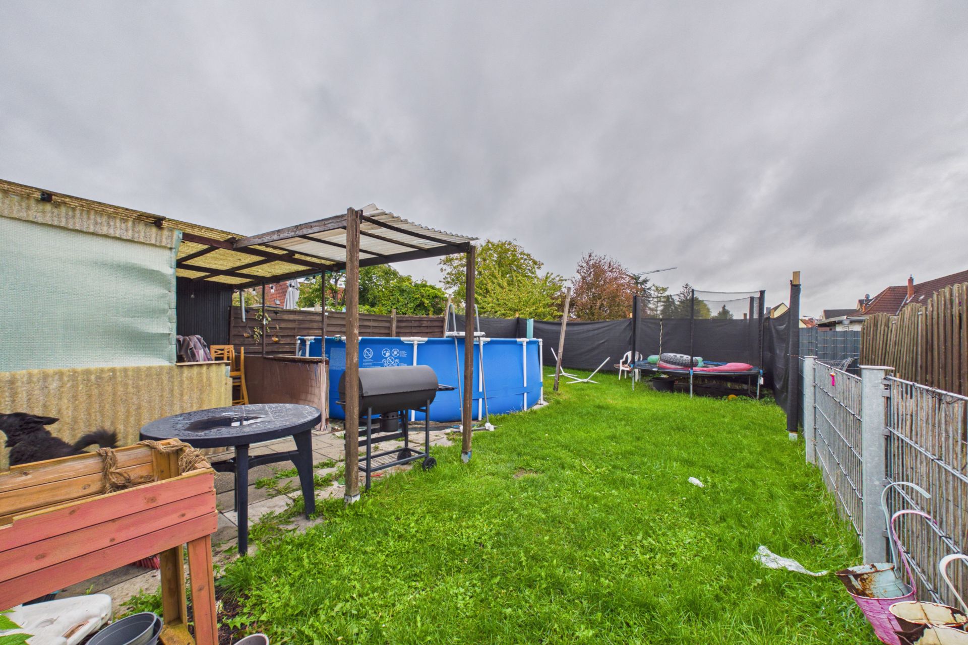 Garten mit Rasen, Grill, Planschbecken, Tisch, Trampolin und überdachtem Bereich.