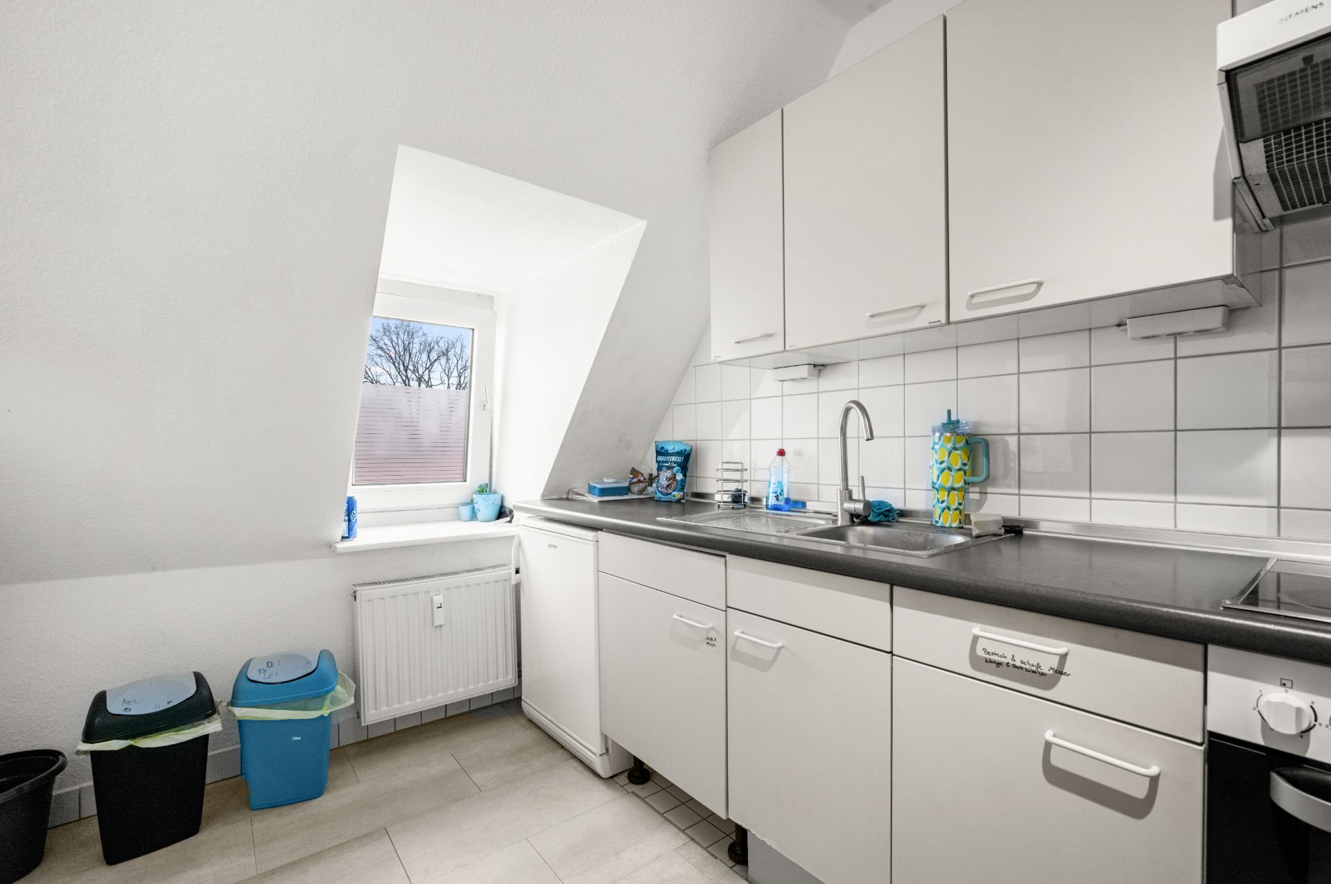 Helle Küche mit weißen Schränken, Spüle, Fenster und Mülleimern an der Wand.
