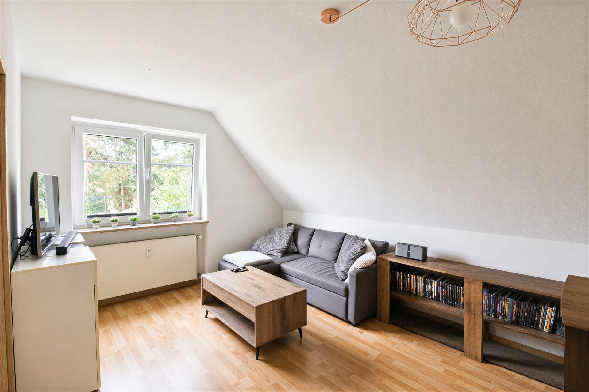 Helles Wohnzimmer mit grauem Sofa, Holztisch, Bücherregal und Fernseher vor einem Fenster.