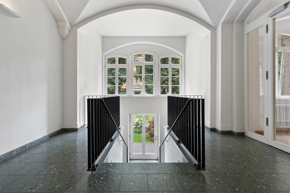 Heller Flur mit großen Fenstern, Treppengeländer und Blick auf Garten mit Blumen.