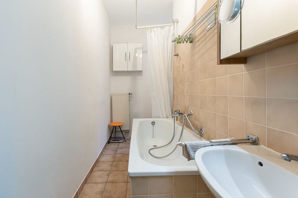 Schmales Badezimmer mit Badewanne, Waschbecken, Spiegel und beigem Fliesenspiegel.