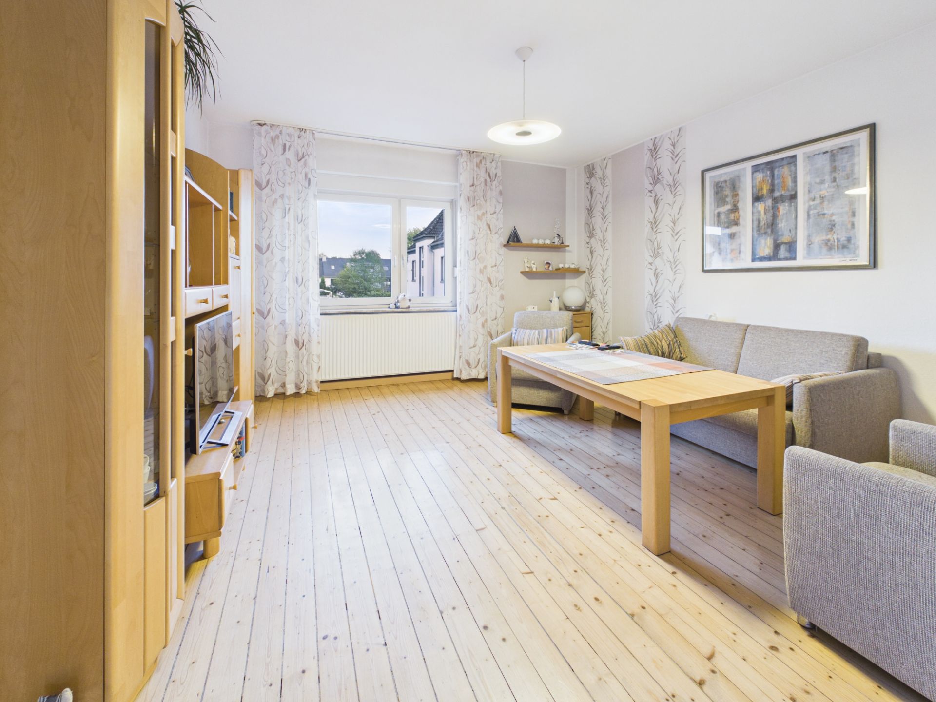 Helles Wohnzimmer mit Holzboden, Sofa, Esstisch, Sessel und großem Fenster.