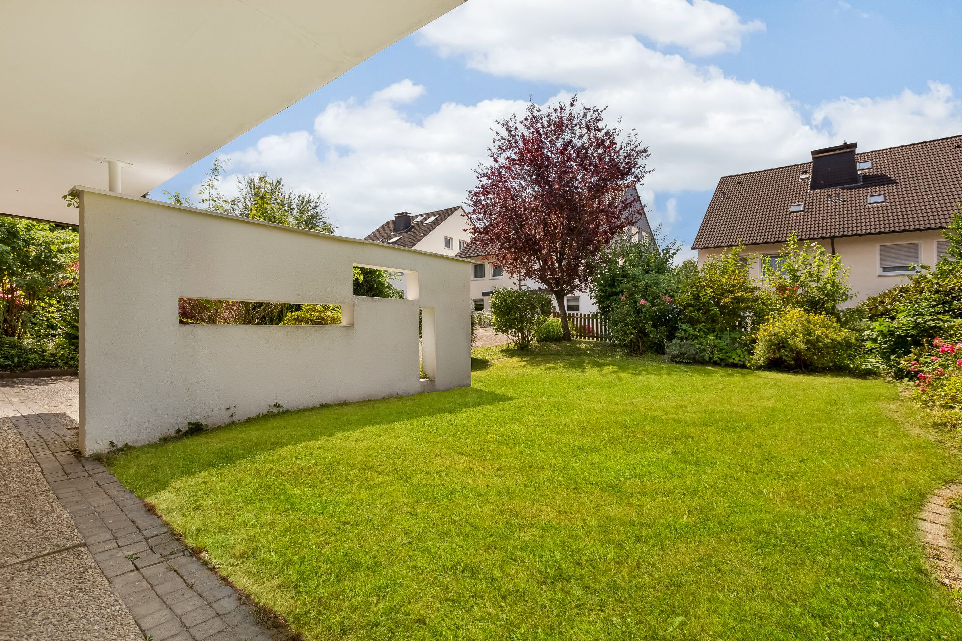 Grüner Garten mit Rasenfläche, Baum, Sträuchern und einer niedrigen weißen Mauer.