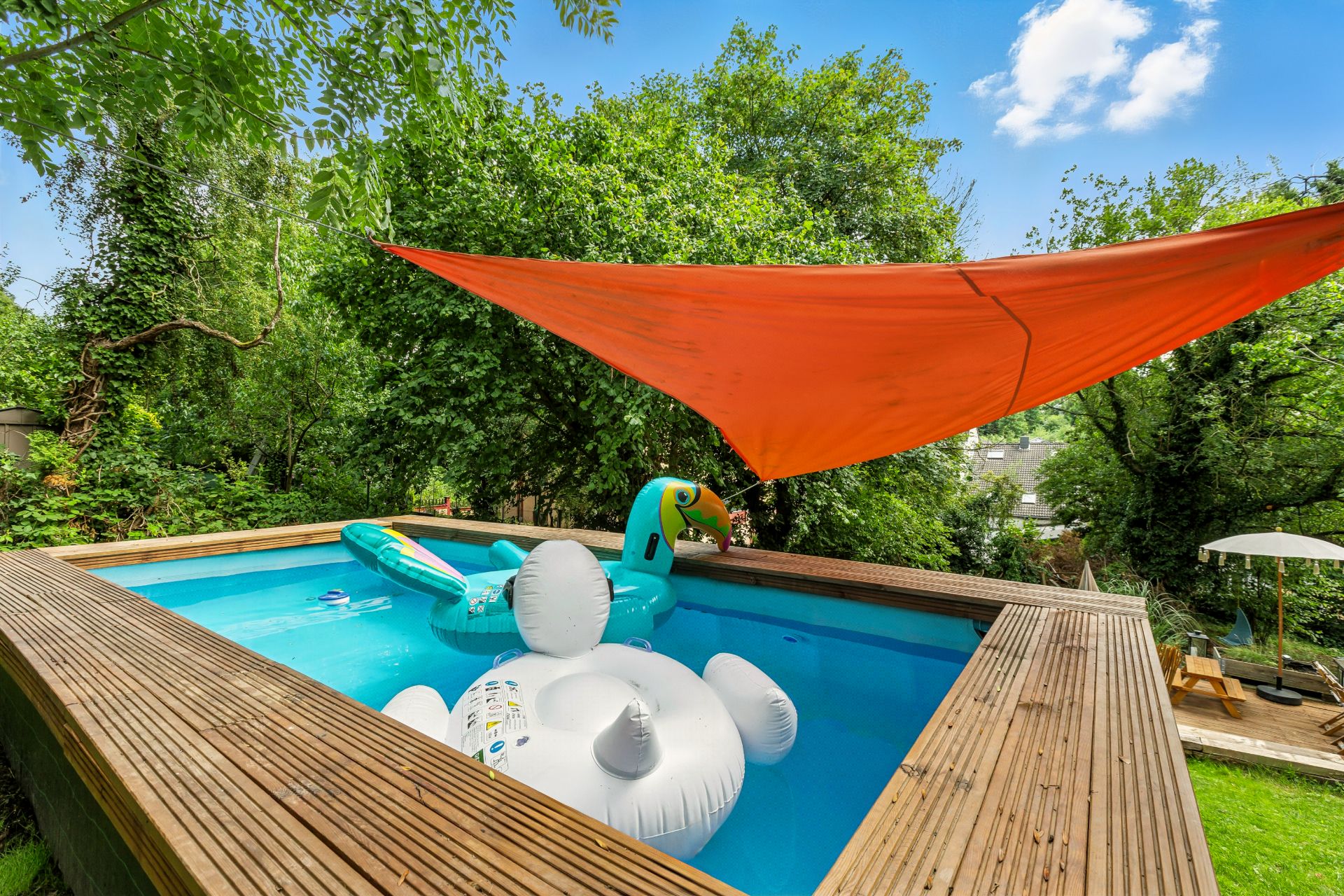Rechteckiger Pool mit Holzumrandung, bunte Schwimmtiere und orangefarbene Markise im Garten.