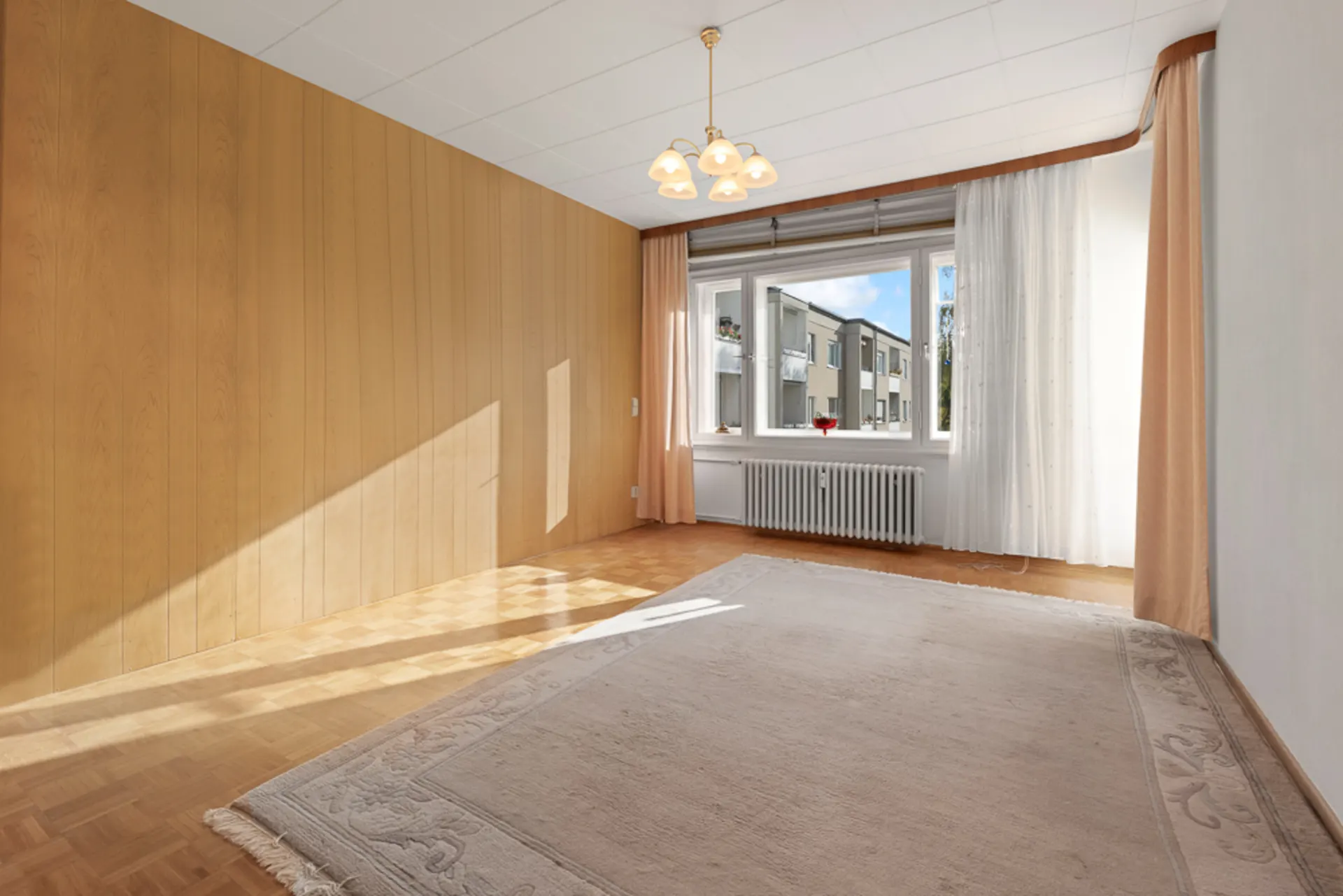 Helles Zimmer mit Holzvertäfelung, großem Fenster, Vorhängen und Teppich auf Holzboden.