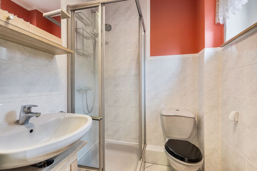 Kleines Bad mit Dusche, Waschbecken und Toilette, weiße Fliesen und rote Wand.