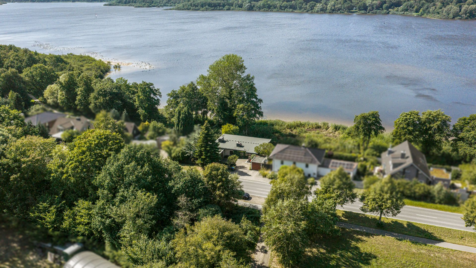 Luftaufnahme von Häusern an einer Straße, mit Bäumen und einem See im Hintergrund.