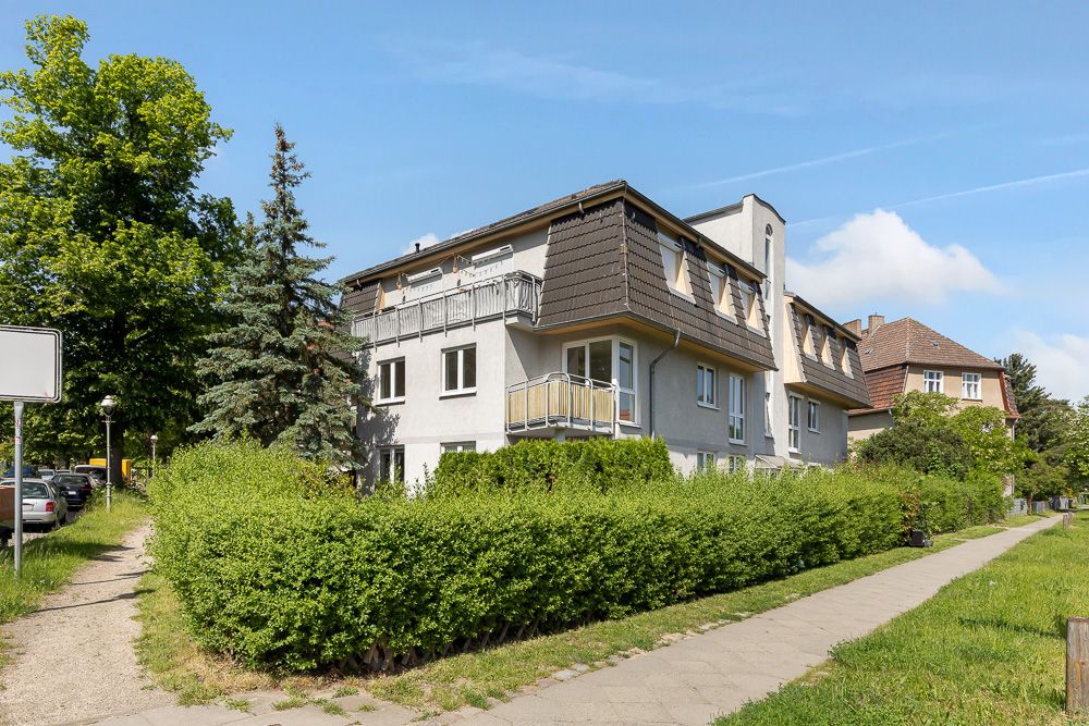 Mehrfamilienhaus mit Balkon, umgeben von Büschen und Bäumen an einer Gehwegkreuzung.