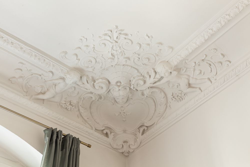 Verzierte Stuckdecke mit Engelmotiven in einer Zimmerecke über einem Fenster mit Vorhang.