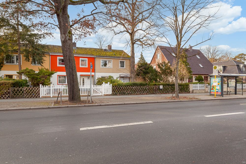 Mehrfamilienhaus mit weißem Zaun und Bushaltestelle davor an einer Straße.