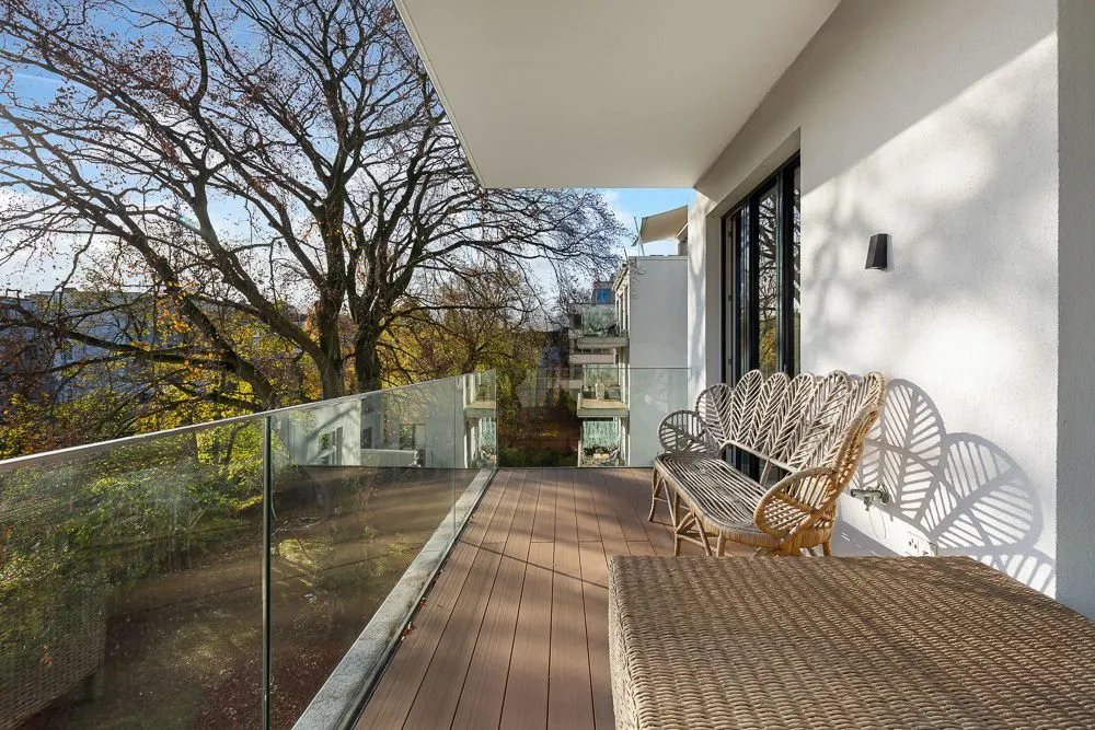 Großer Balkon mit Glasgeländer, Rattanmöbeln und Blick auf Bäume.