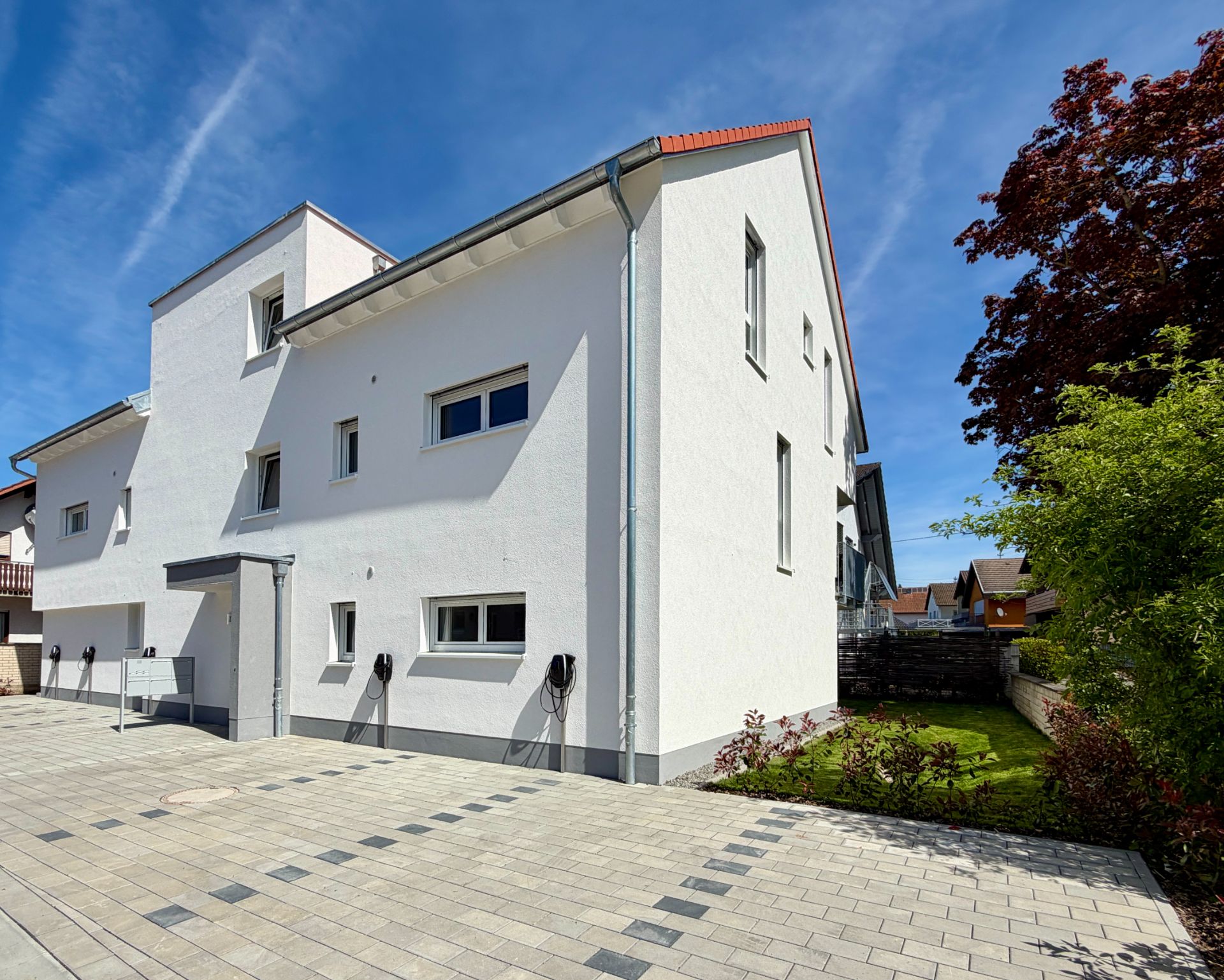 Modernes, weißes Wohnhaus mit Flachdach und gepflastertem Hof, daneben ein kleiner Garten mit Bäumen.