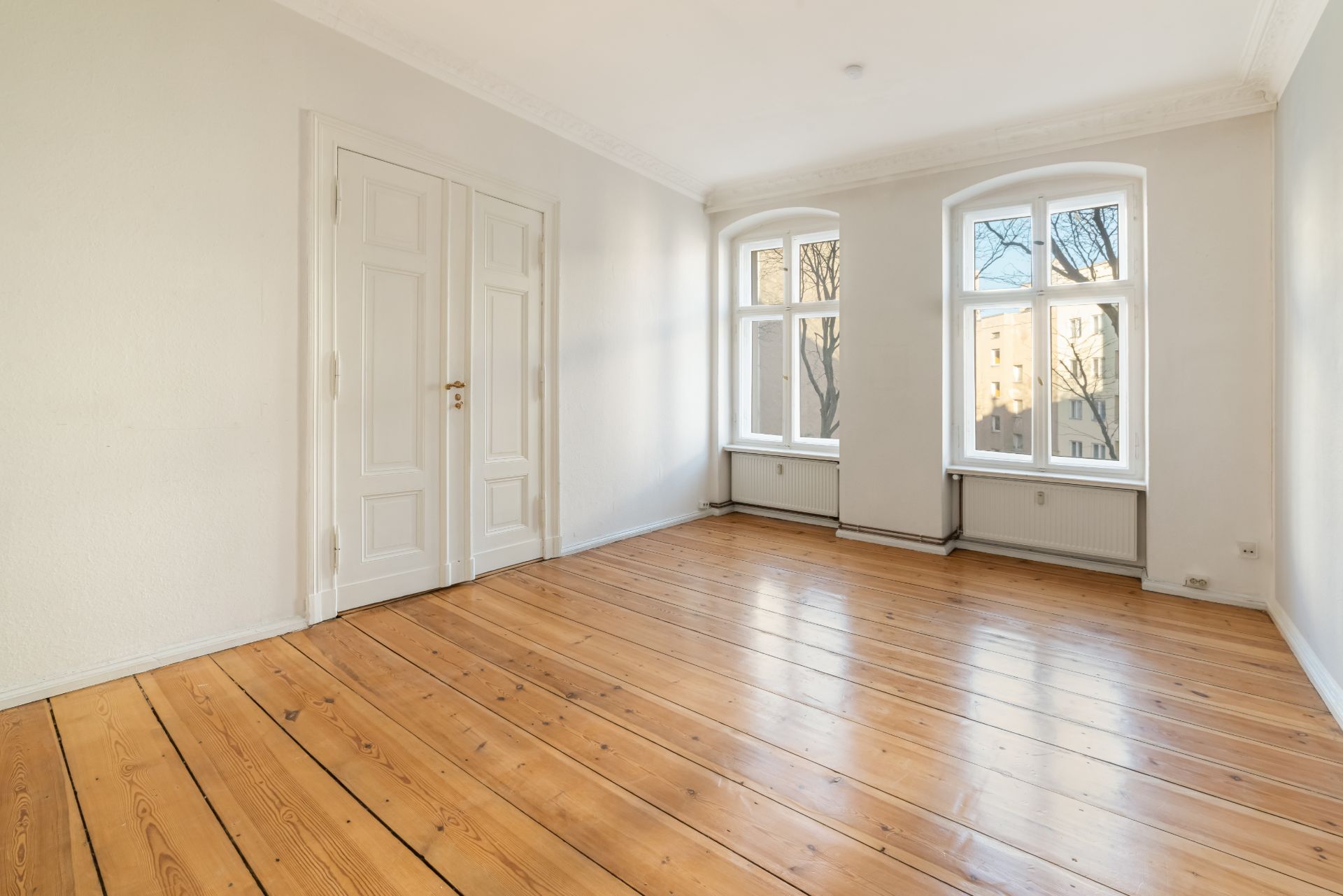 Helles, leeres Zimmer mit Holzboden, weißen Wänden, Doppeltür und zwei großen Fenstern.