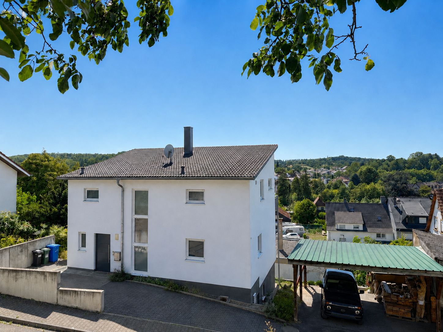 Weißes Wohnhaus mit Flachdach und Carport, umgeben von weiteren Häusern und Bäumen.