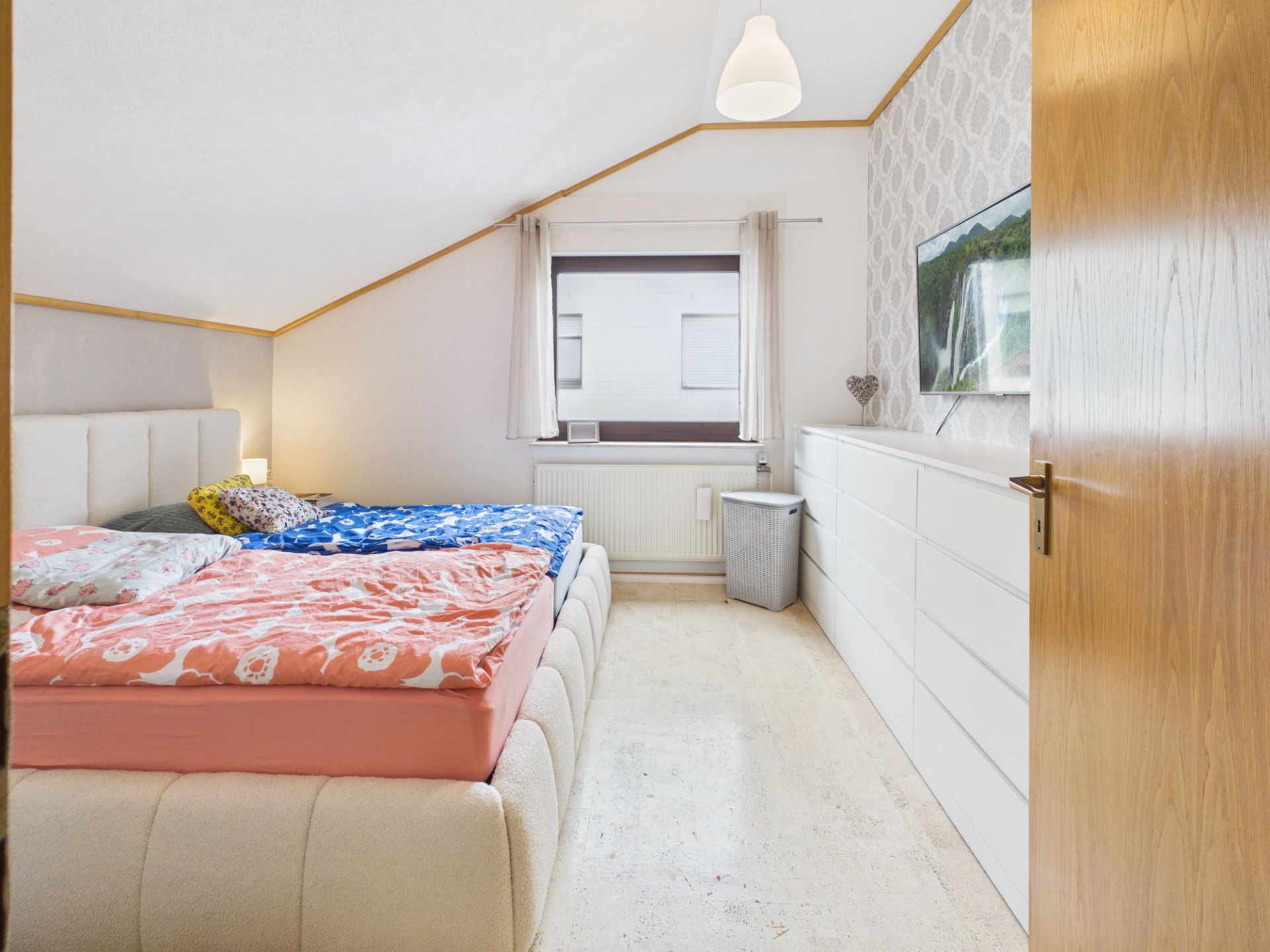Helles Schlafzimmer mit Doppelbett, weißer Kommode und Fenster unter einer Dachschräge.