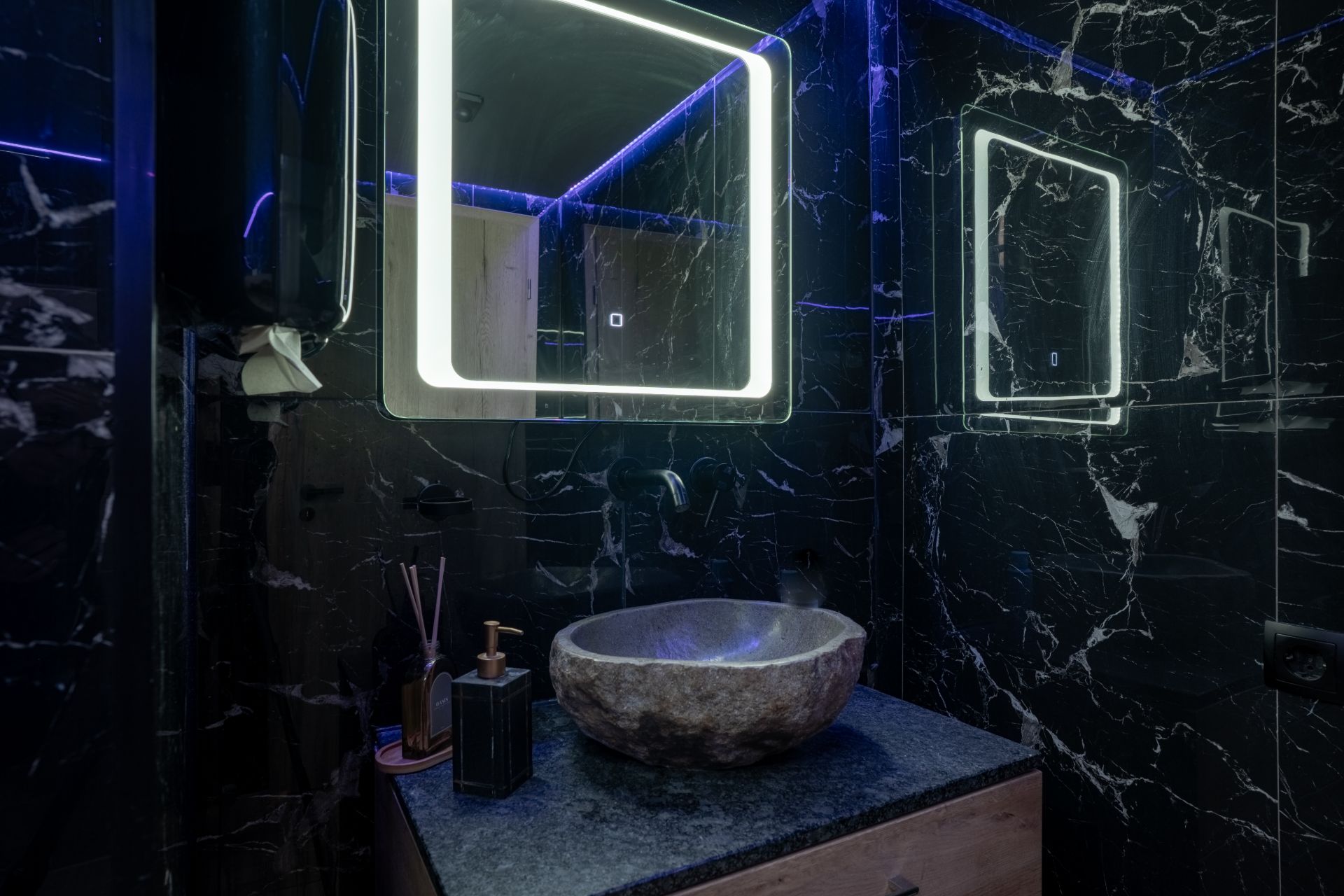 Modernes Badezimmer mit beleuchtetem Spiegel und rundem Steinauswaschbecken auf dunkler Marmorplatte.