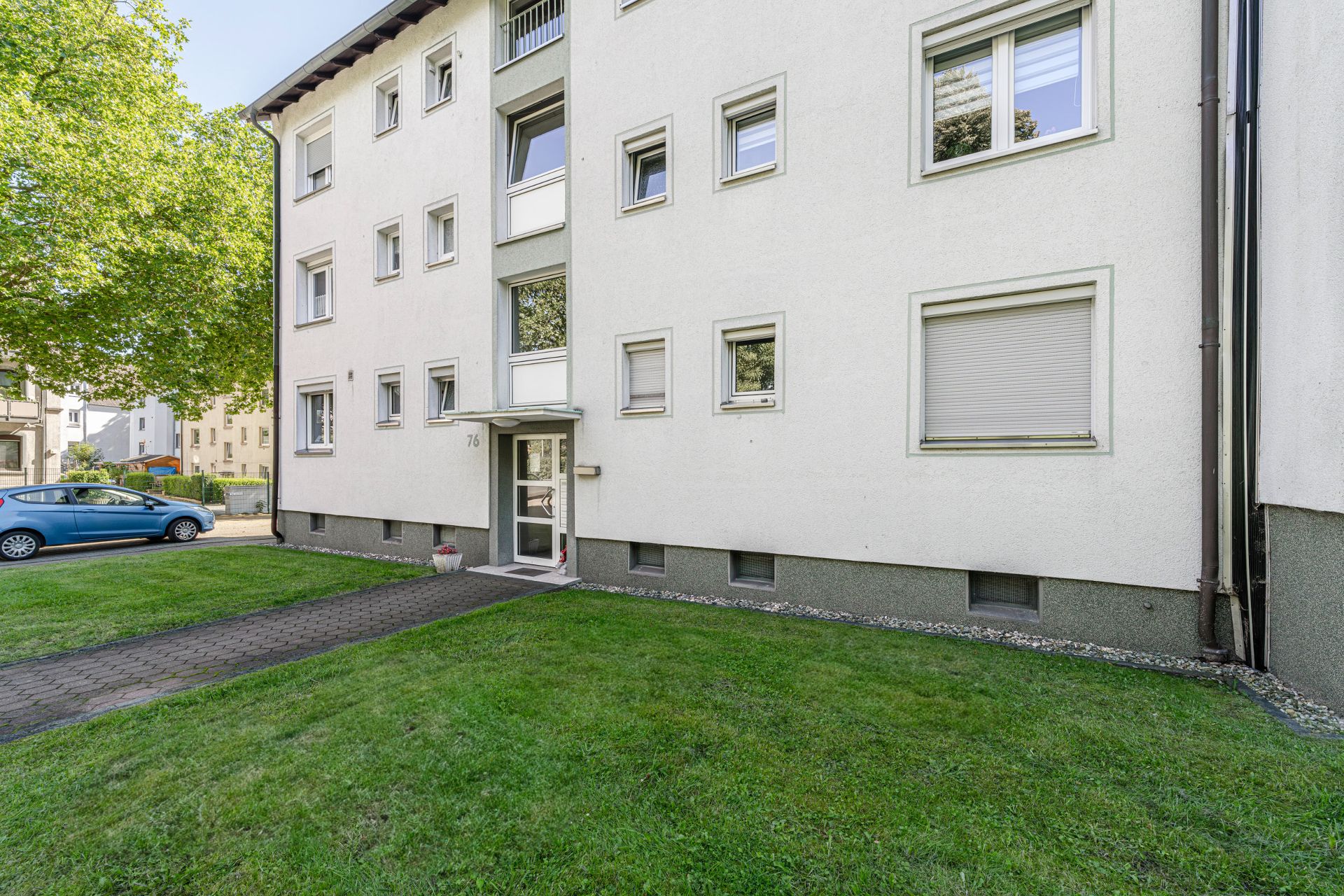 Mehrfamilienhaus mit hellem Putz, mehreren Fenstern und Eingangstür, davor gepflegter Rasen.