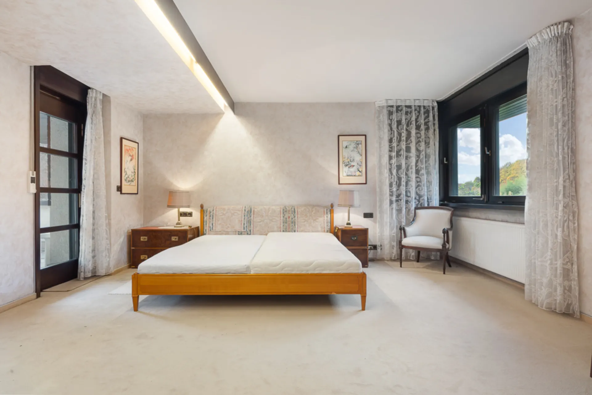 Helles Schlafzimmer mit Doppelbett, zwei Nachttischen, Stühlen und großen Fenstern mit Vorhängen.