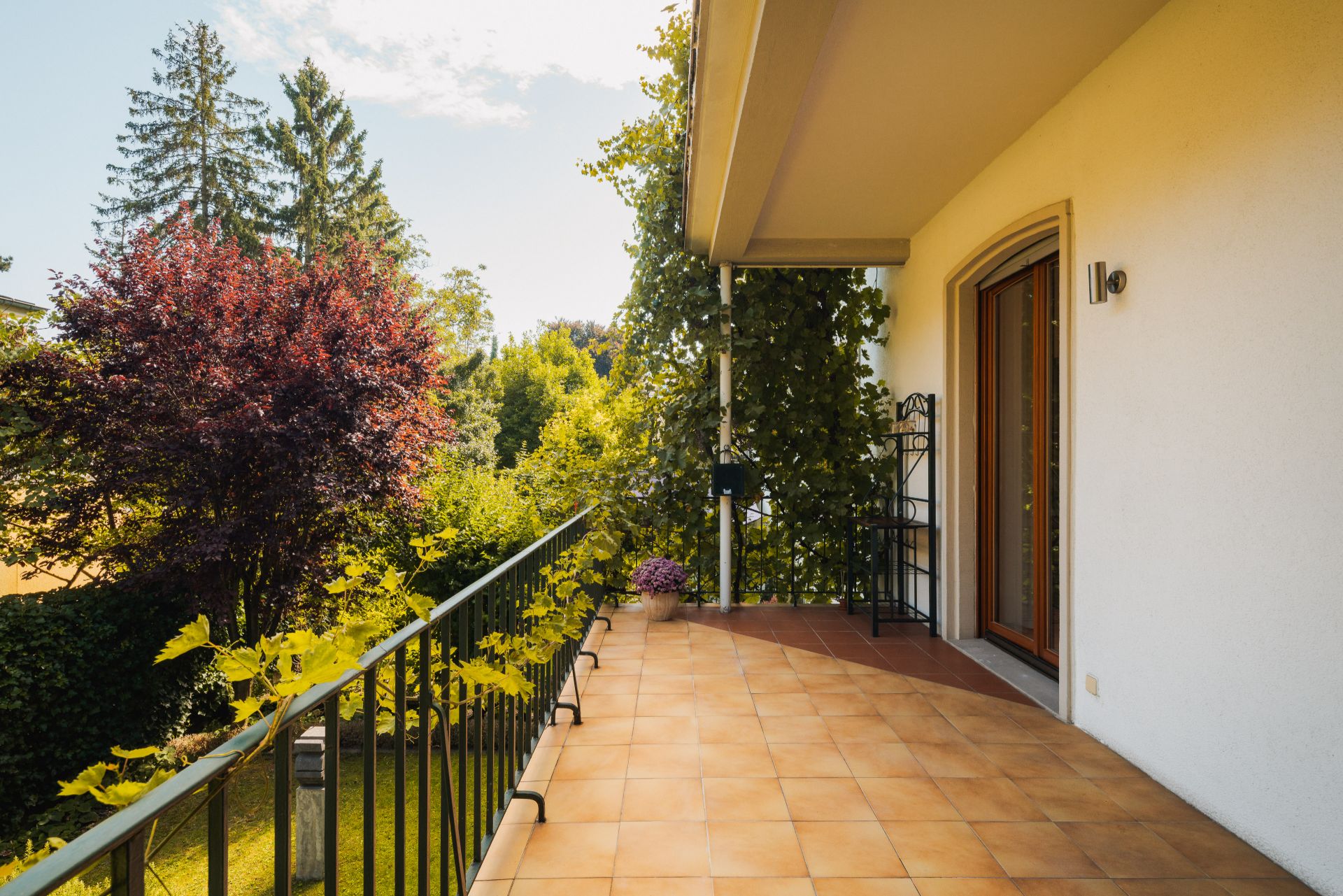 Großer Balkon mit Fliesenboden, Pflanzen am Geländer und Blick auf Garten mit Bäumen.