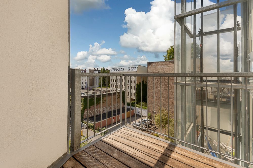 Kleiner Balkon mit Holzboden und Metallgeländer, Blick auf Nachbargebäude und Himmel mit Wolken.