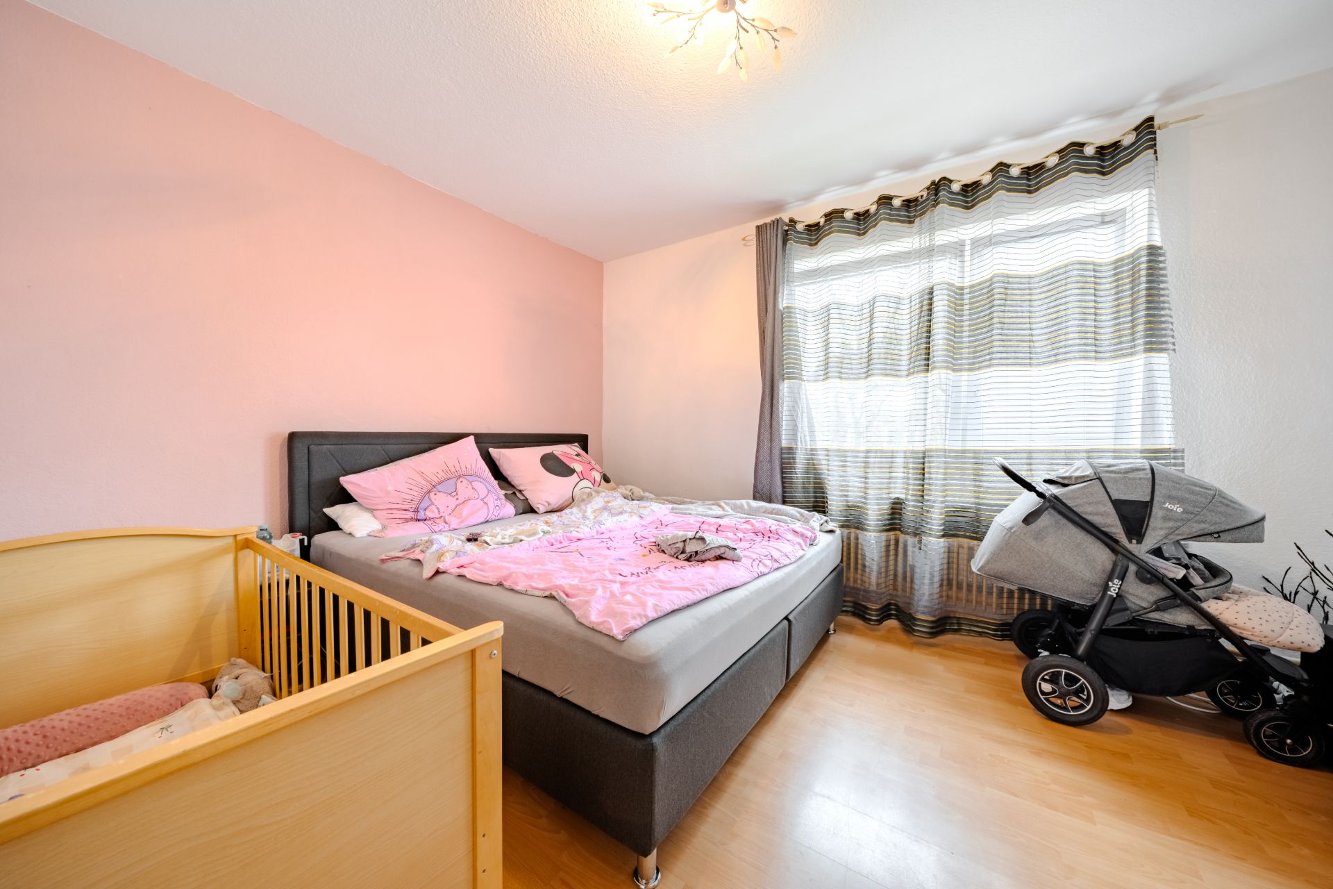 Schlafzimmer mit Doppelbett, Babybett, Kinderwagen und großem Fenster mit Vorhängen.
