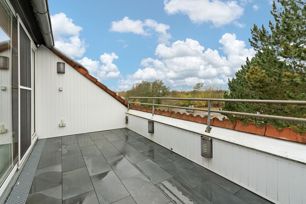 Große Dachterrasse mit grauen Fliesen, weißer Wand und Metallgeländer, Blick ins Grüne.
