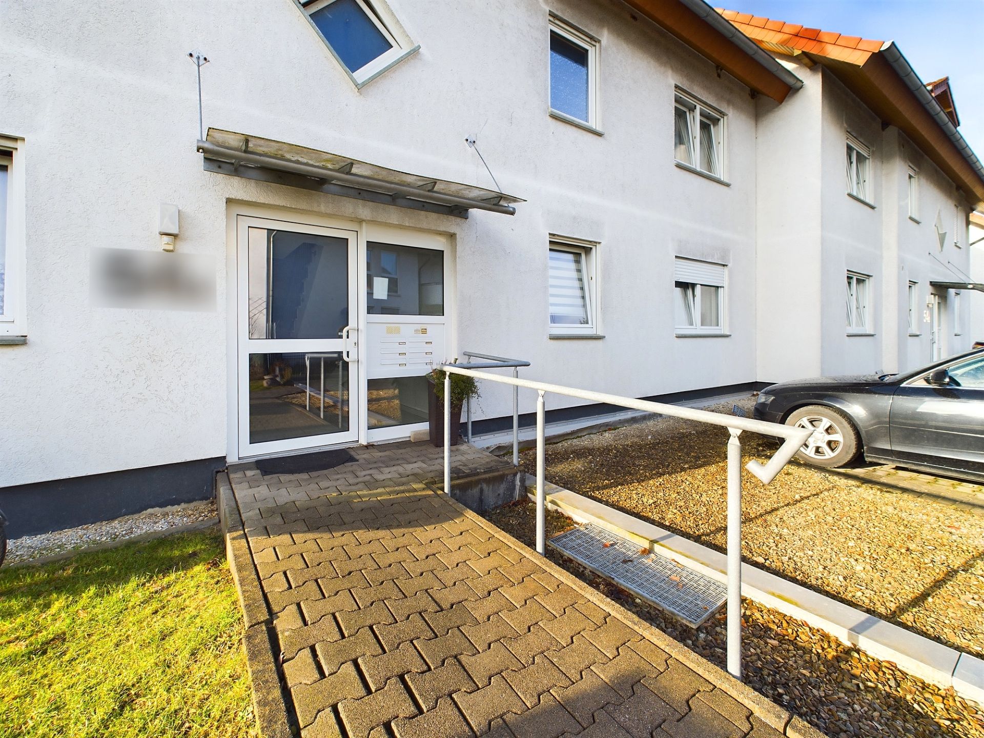 Eingang eines Wohnhauses mit Rampe, Glasdach und geparktem Auto rechts.